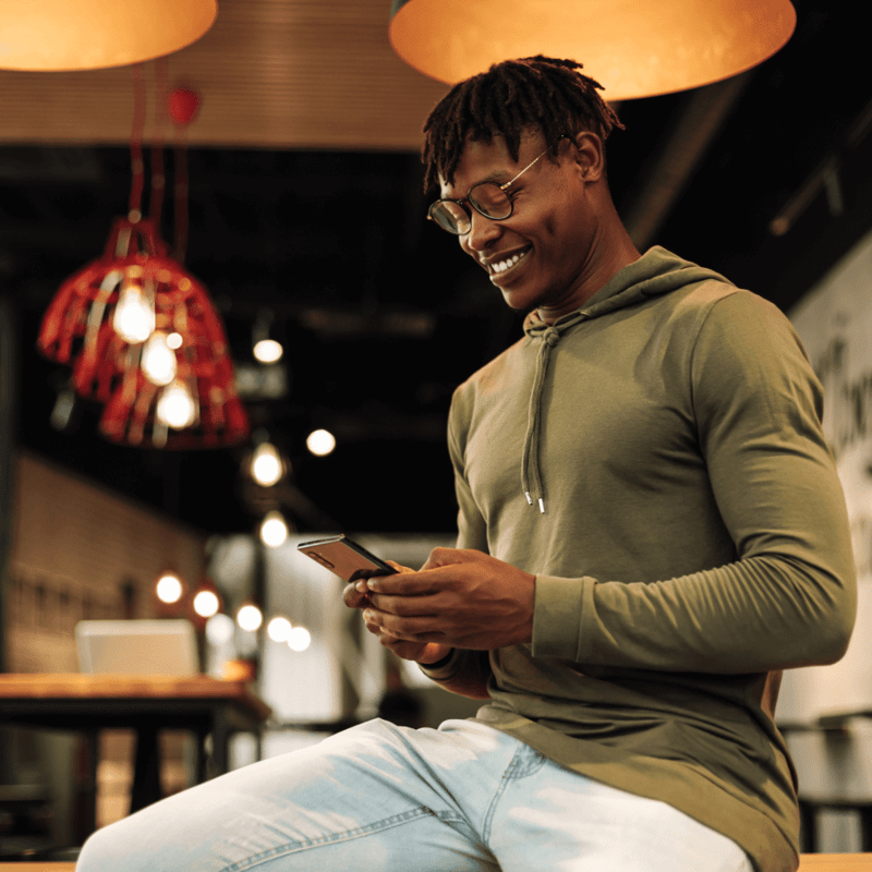 Man wearing glasses and a green hoodie in a cafe looking at mobile phone in his hands